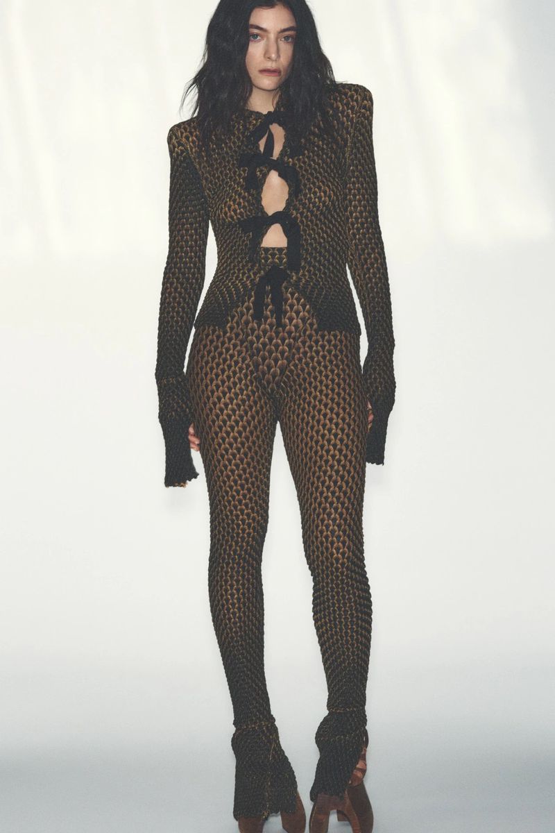 A woman with long, dark curly hair poses in a full-length shot against a white background, wearing a lace-up top and leggings.