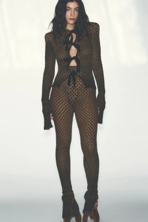 A woman with long, dark curly hair poses in a full-length shot against a white background, wearing a lace-up top and leggings.