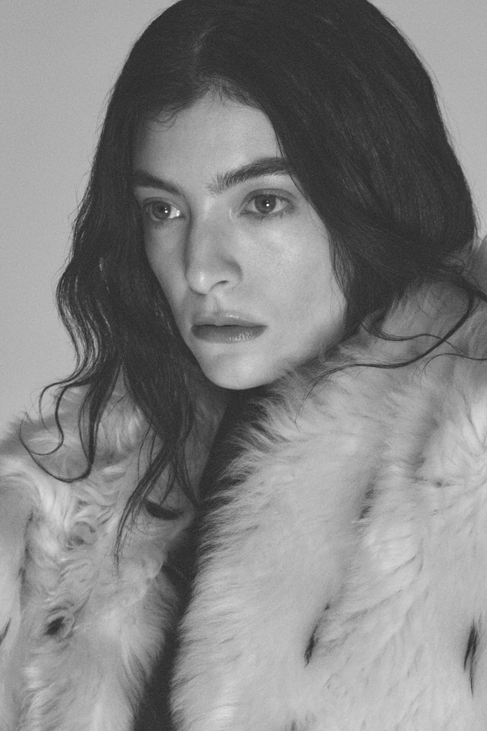 Portrait of singer Lorde with dark hair wearing a fluffy textured coat against a plain background
