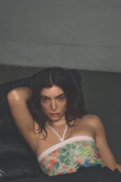 Lorde in a floral patterned top leaning against a dark surface in a minimalist interior setting.
