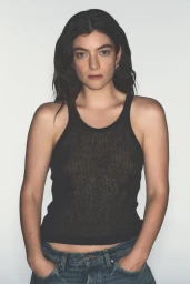Musician Lorde wearing a sheer black ribbed tank top and blue jeans with hands in pockets against a plain white studio backdrop.
