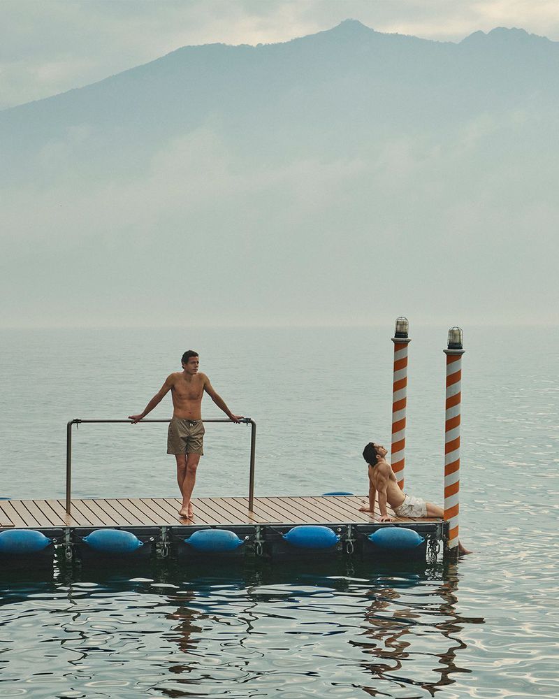 Two male models in swim shorts posing on a wooden floating dock on a calm lake with distant hazy mountains.