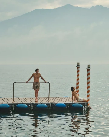 Two male models in swim shorts posing on a wooden floating dock on a calm lake with distant hazy mountains.