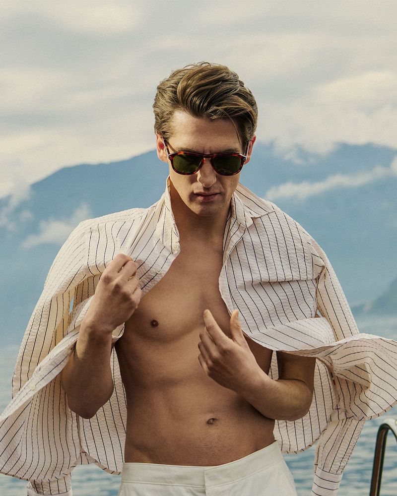 Man wearing sunglasses and a striped shirt outdoors against a scenic mountain lake background