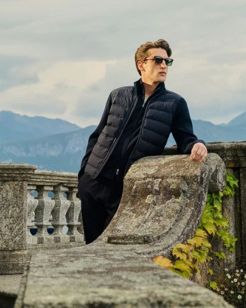 A man wearing sunglasses and a navy quilted vest leans against a stone balustrade overlooking misty mountains.