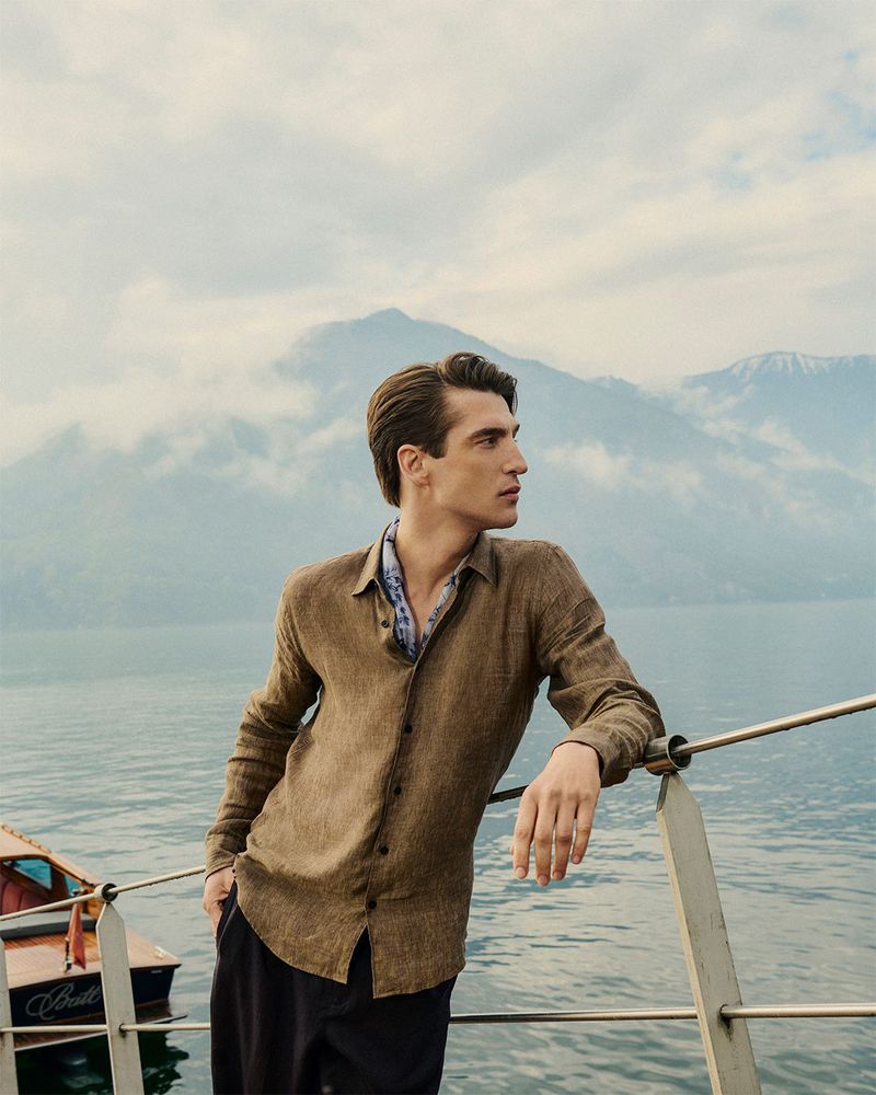 Man in a brown linen shirt and neckerchief leaning against a boat railing with a lake and mountains behind him.