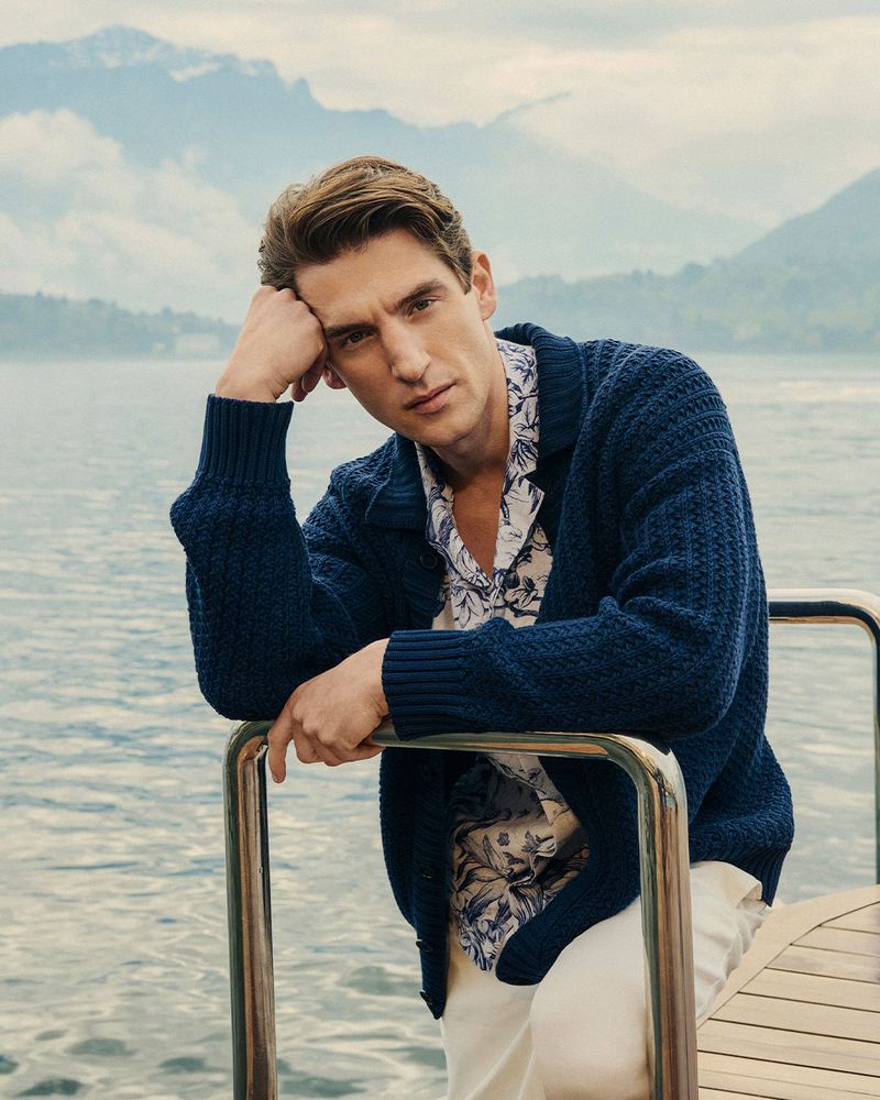 Anatol Modzelewski wearing a navy knit cardigan and floral print shirt on a boat with a lake and mountains in the background.