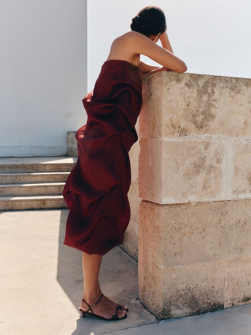 Model Charlotte Koepf in a strapless burgundy draped dress and kitten-heel sandals leaning against a stone pillar outdoors.
