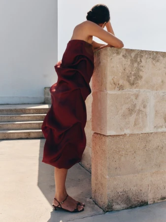 Model Charlotte Koepf in a strapless burgundy draped dress and kitten-heel sandals leaning against a stone pillar outdoors.