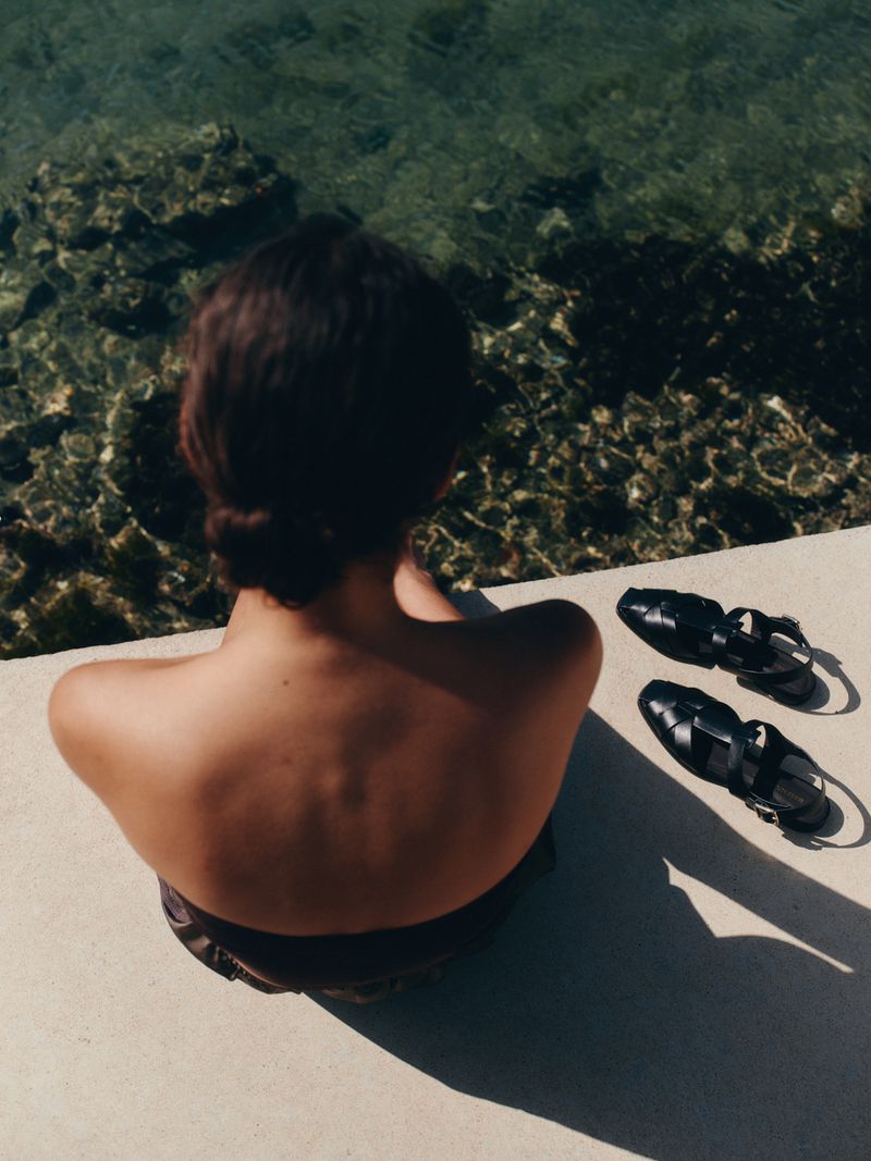 High-angle rear view of a woman seated on a stone ledge next to a pair of black leather sandals above clear coastal water.