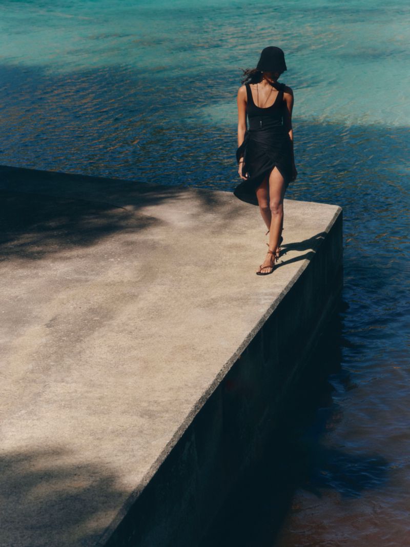 Woman in a black tank dress and black bucket hat walking along the edge of a concrete pier overlooking turquoise water.