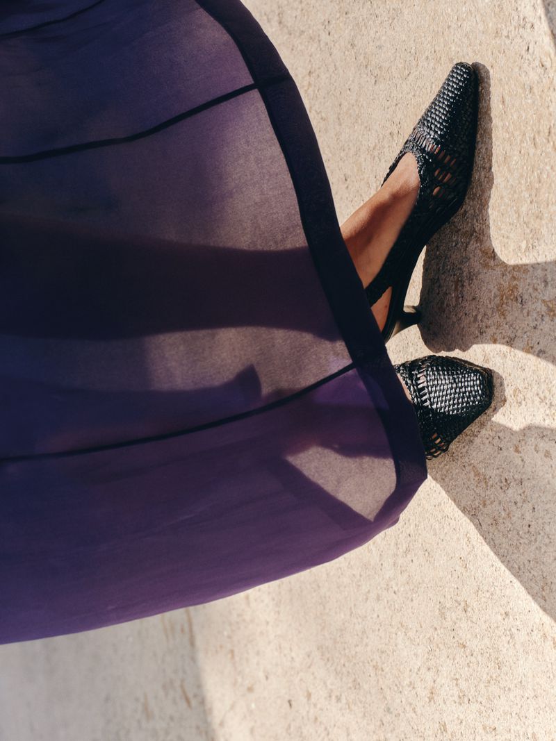 Model wearing woven black slingback kitten heels and a sheer purple maxi dress on a sunlit stone ground.