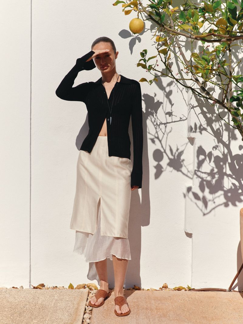 Model Charlotte Koepf standing by a white stucco wall and lemon tree wearing a black cardigan and cream skirt.