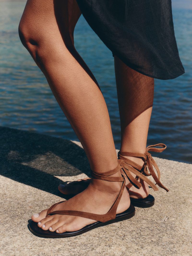 Close-up of a model's feet wearing brown suede lace-up flat sandals while standing on a stone ledge next to the water.