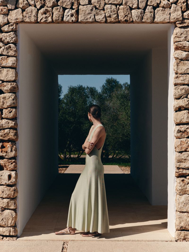 Model wearing a light green sleeveless long dress standing inside a stone archway looking towards trees outdoors