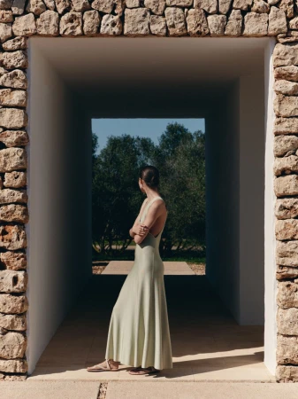 Model wearing a light green sleeveless long dress standing inside a stone archway looking towards trees outdoors