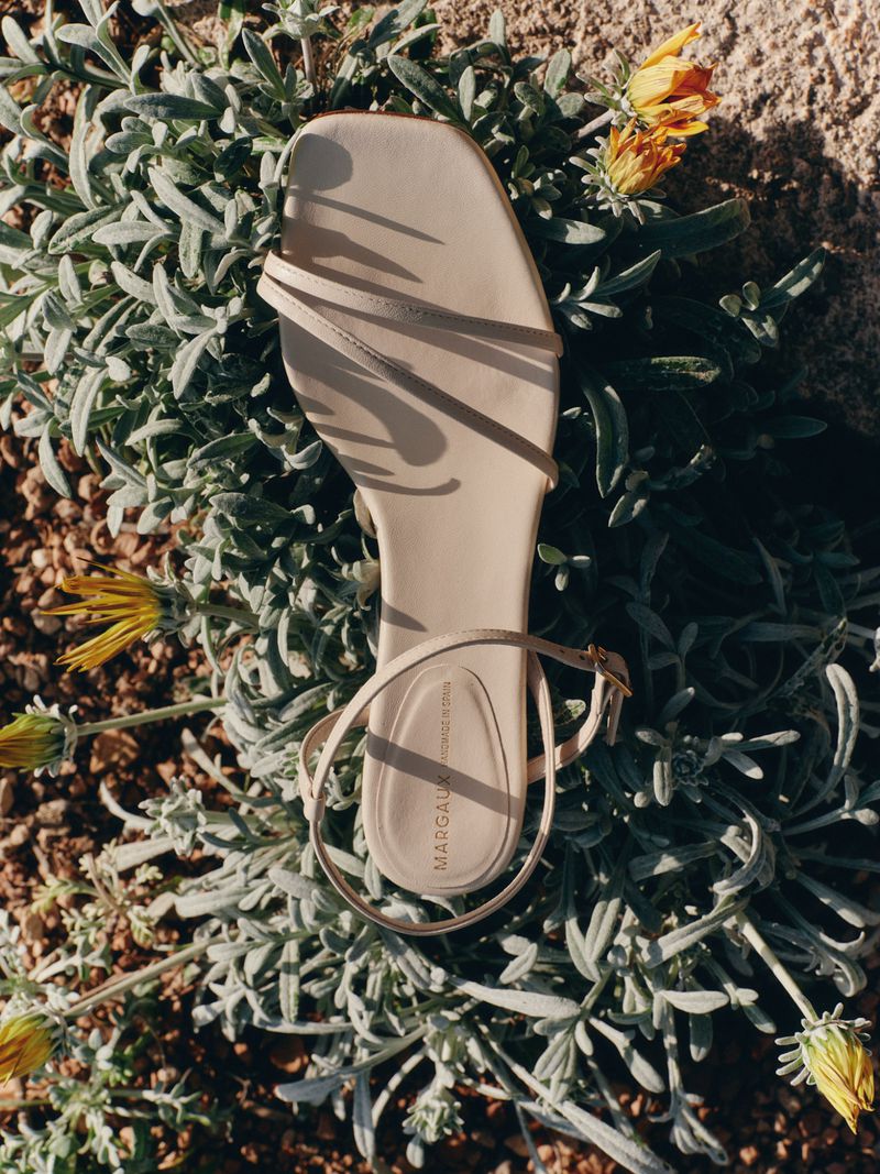 Beige strappy flat sandal with a square toe resting on green foliage with small yellow flowers in direct sunlight.