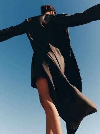 Low-angle view of model Charlotte Koepf in a flowing black coat with arms outstretched against a clear blue sky.