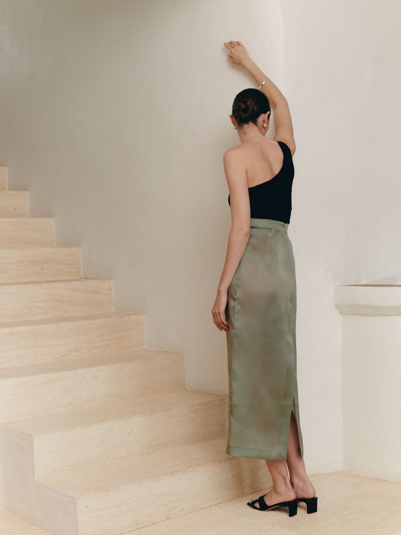 Model standing on a pale staircase wearing a black top and a green satin skirt with one arm raised against the wall.