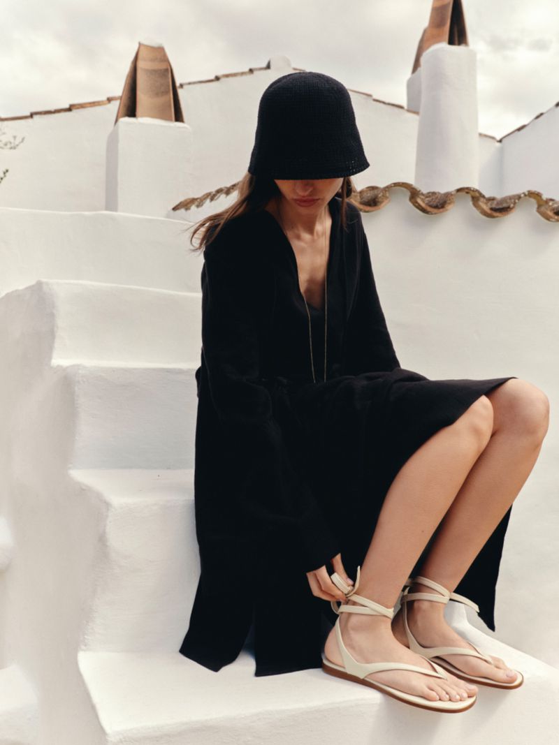 Model Charlotte Koepf in a black dress and cloche hat fastening flat cream wrap sandals while seated on white stucco steps.