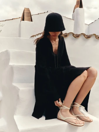 Model Charlotte Koepf in a black dress and cloche hat fastening flat cream wrap sandals while seated on white stucco steps.