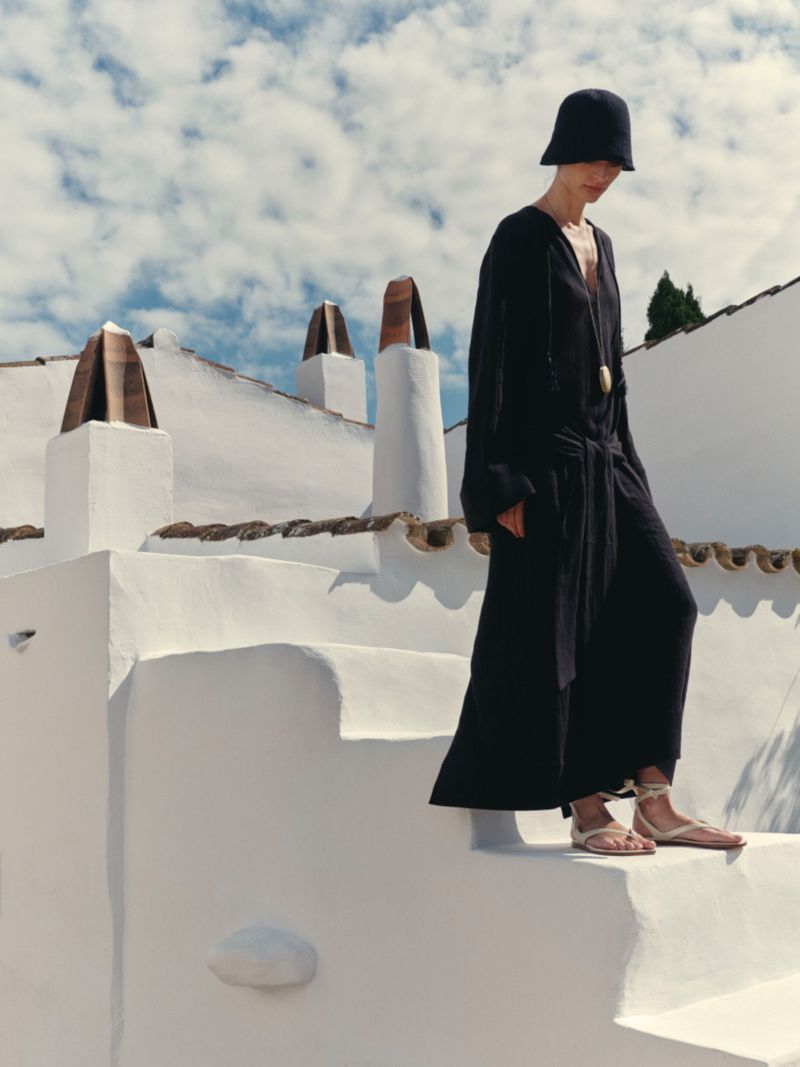 Model Charlotte Koepf wearing a black maxi dress, black hat, and lace-up flat sandals on whitewashed Mediterranean stairs.
