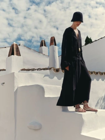 Model Charlotte Koepf wearing a black maxi dress, black hat, and lace-up flat sandals on whitewashed Mediterranean stairs.