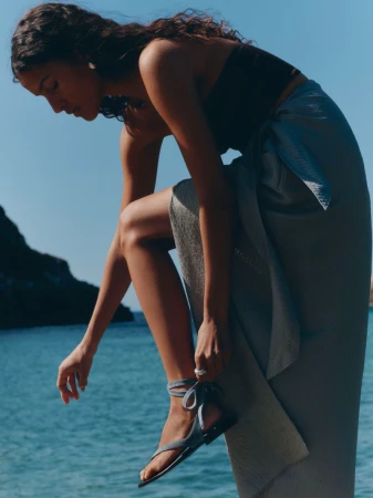 Charlotte Koepf adjusting an ankle-strap flat sandal wearing a wrap skirt by the sea for Margaux NY.