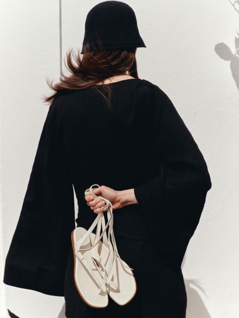 Back view of a woman wearing a black bell-sleeve dress and black knit hat holding white strappy flat sandals against a sunlit white wall.