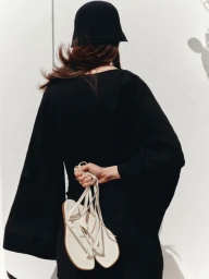 Back view of a woman wearing a black bell-sleeve dress and black knit hat holding white strappy flat sandals against a sunlit white wall.