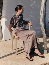 Model Charlotte Koepf in a taupe satin midi dress and black strappy sandals seated outdoors on a woven chair in sunlight.