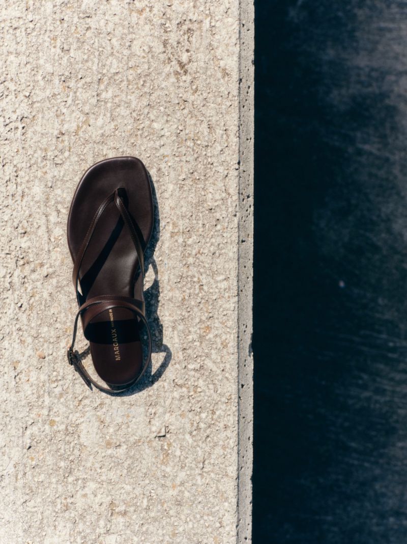Dark brown leather strap sandal resting on a textured light stone ledge beside deep water.