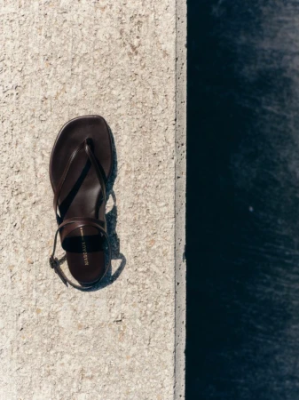Dark brown leather strap sandal resting on a textured light stone ledge beside deep water.