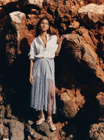 Charlotte Koepf posing against a red rock wall in an unbuttoned white shirt, pale blue fringe skirt, and white strappy sandals.