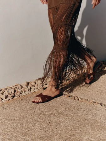Close-up of a model walking in brown cross-strap flat slide sandals and a long brown fringed skirt along a sunny concrete walkway.