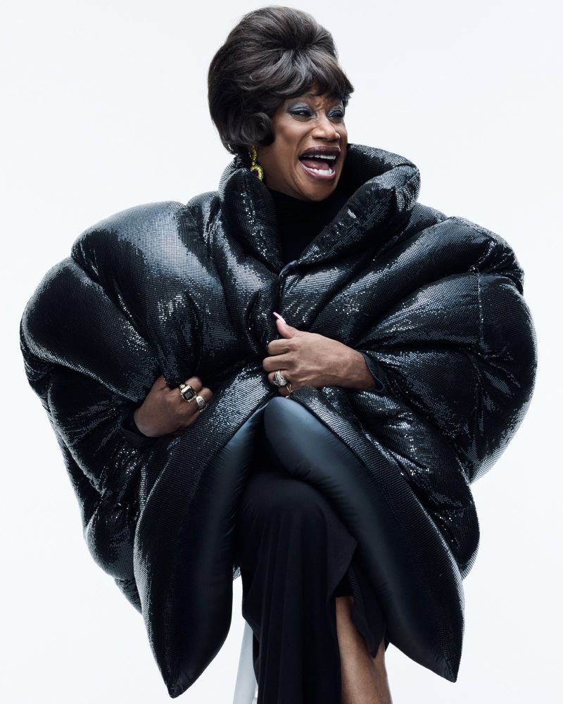 Person in a massive black sequined puffer coat with a retro styled bouffant wig smiling against a white studio backdrop.