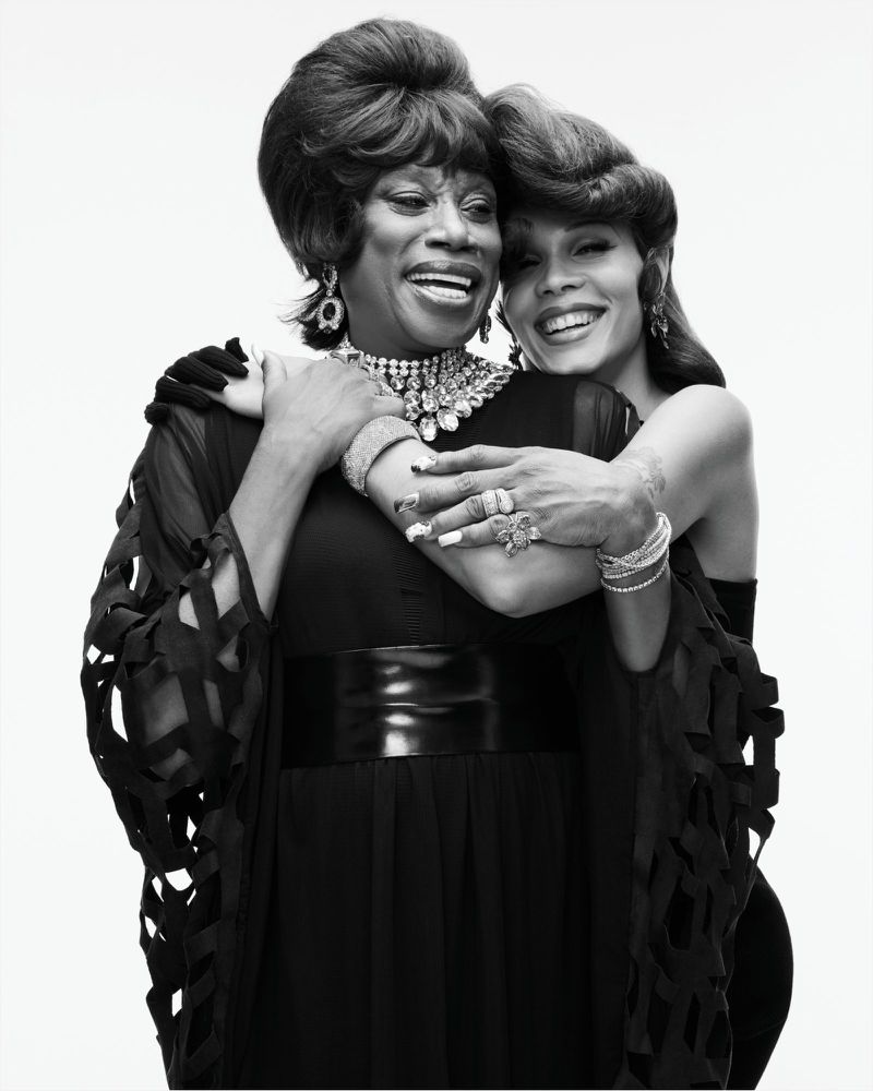 Black and white studio portrait of two smiling people embracing, styled with retro bouffant hair and ornate statement jewelry.