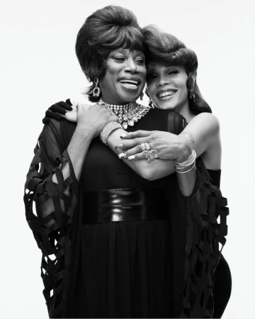Black and white studio portrait of two smiling people embracing, styled with retro bouffant hair and ornate statement jewelry.