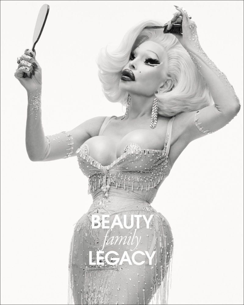 Black and white portrait of Amanda Lepore holding a hand mirror and comb in an embellished corset dress against a white studio background.