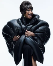 Person in a massive black sequined puffer coat with a retro styled bouffant wig smiling against a white studio backdrop.
