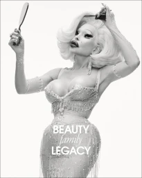 Black and white portrait of Amanda Lepore holding a hand mirror and comb in an embellished corset dress against a white studio background.