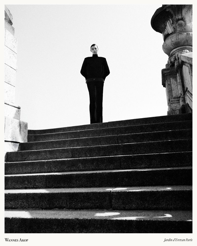 Model standing at the top of a grand stone staircase wearing dark fitted apparel under a bright sky.