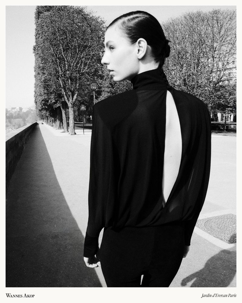 Model wearing a black draped backless top viewed from behind on an outdoor tree-lined walkway in black and white.