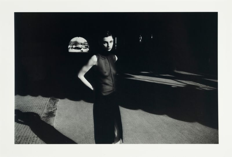 Model Estella Nolan standing in a dark architectural walkway illuminated by sharp geometric sunlight and shadow patterns.
