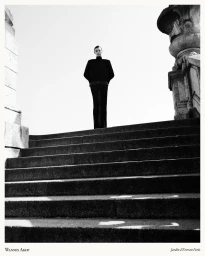 Model standing at the top of a grand stone staircase wearing dark fitted apparel under a bright sky.