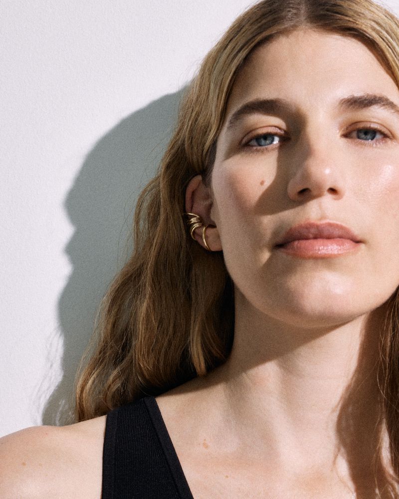 Close-up portrait of a woman wearing gold ear cuffs and a black top against a plain white wall with sharp shadows.