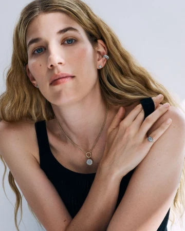 Veronika Heilbrunner wearing a gold pendant necklace, ear cuff, and black tank top against a neutral studio backdrop.