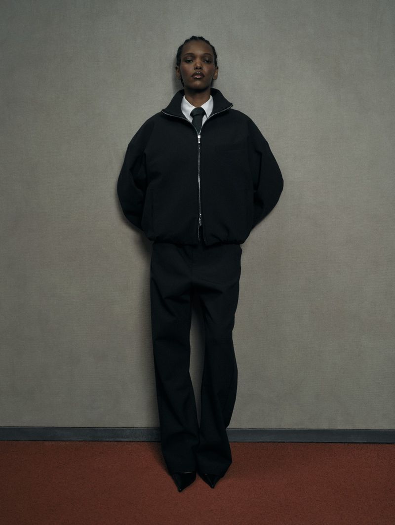 Model Nicole Atieno poses against a grey wall in an oversized black zip jacket, wide trousers, and a white shirt with a black tie.