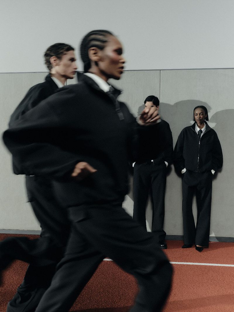 Models in dark clothing walking past a wall with other figures standing against it