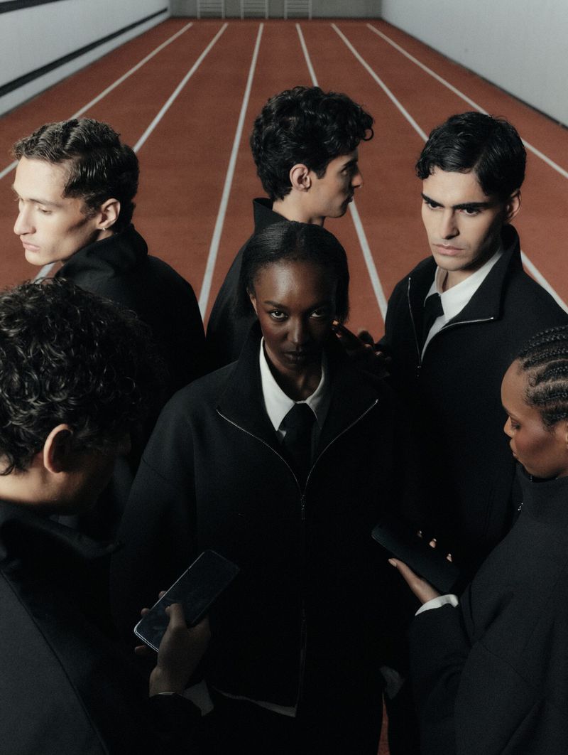 Nicole Atieno and models wearing black zip jackets and ties standing close together on a red running track.